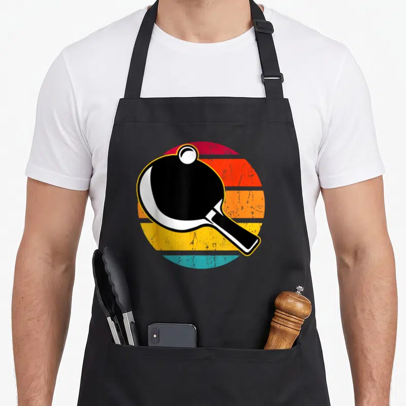 Tablier de cuisine noir avec illustration rétro d'une raquette de ping pong et une balle blanche sur un cercle aux couleurs vives.