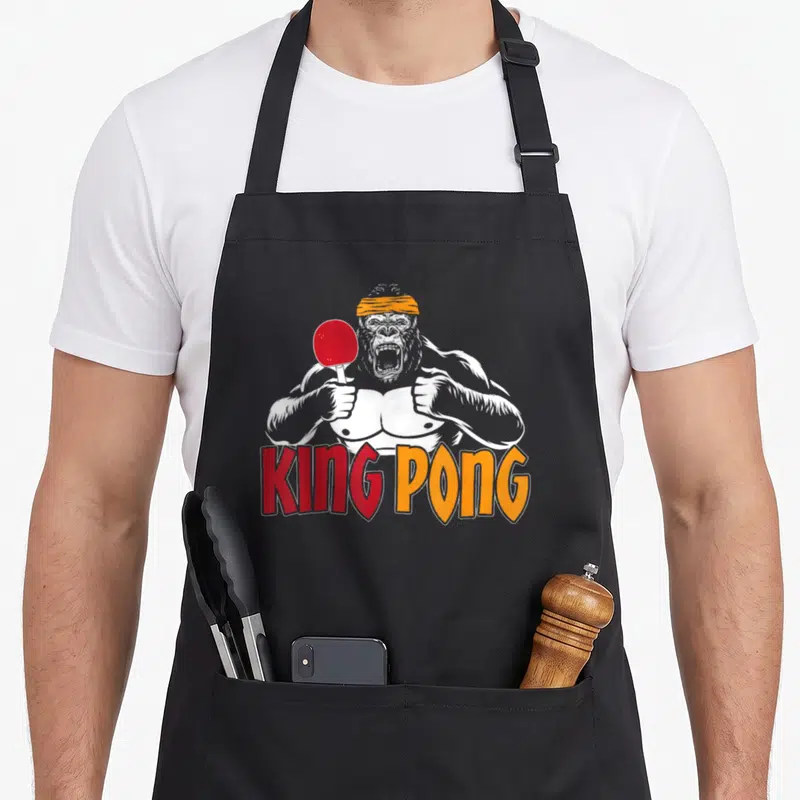 Tablier de cuisine noir avec un gorille musclé tenant une raquette rouge et le texte King Pong en lettres colorées.