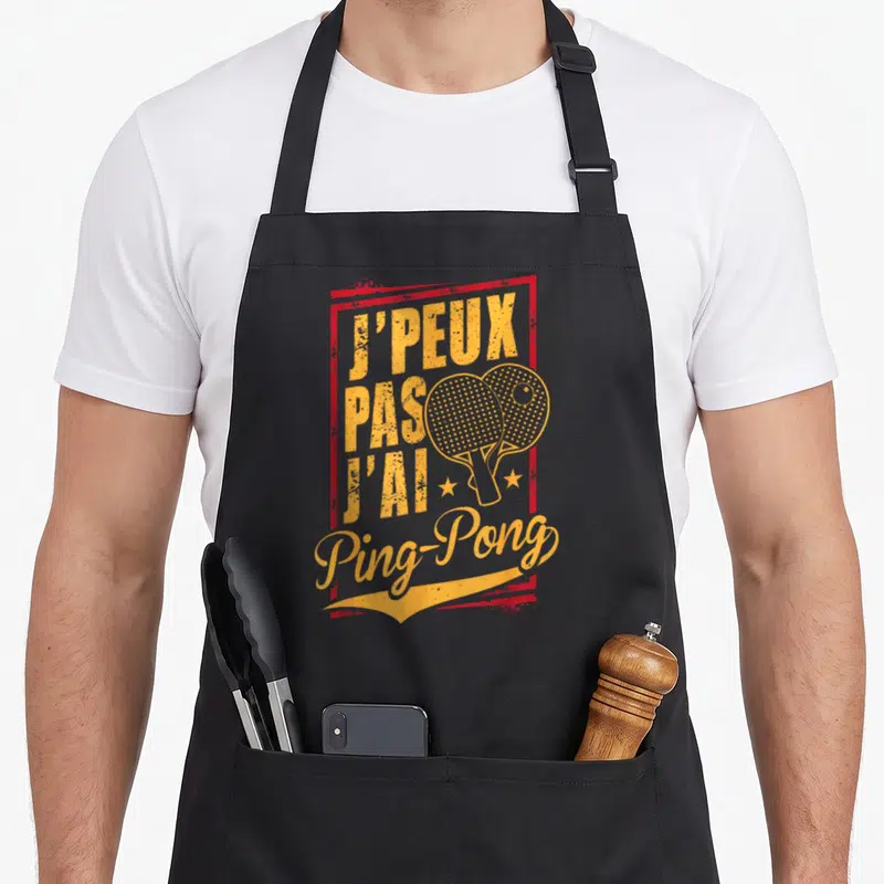 Tablier de cuisine noir avec inscription J'peux pas j'ai Ping-Pong et deux raquettes de ping-pong jaunes.