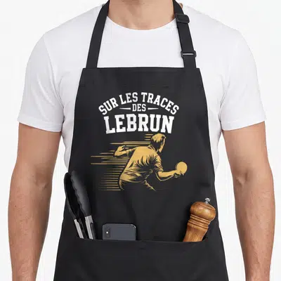 Tablier de cuisine noir avec illustration jaune d'un joueur de ping-pong et le texte Sur les traces des Lebrun en typographie blanche.