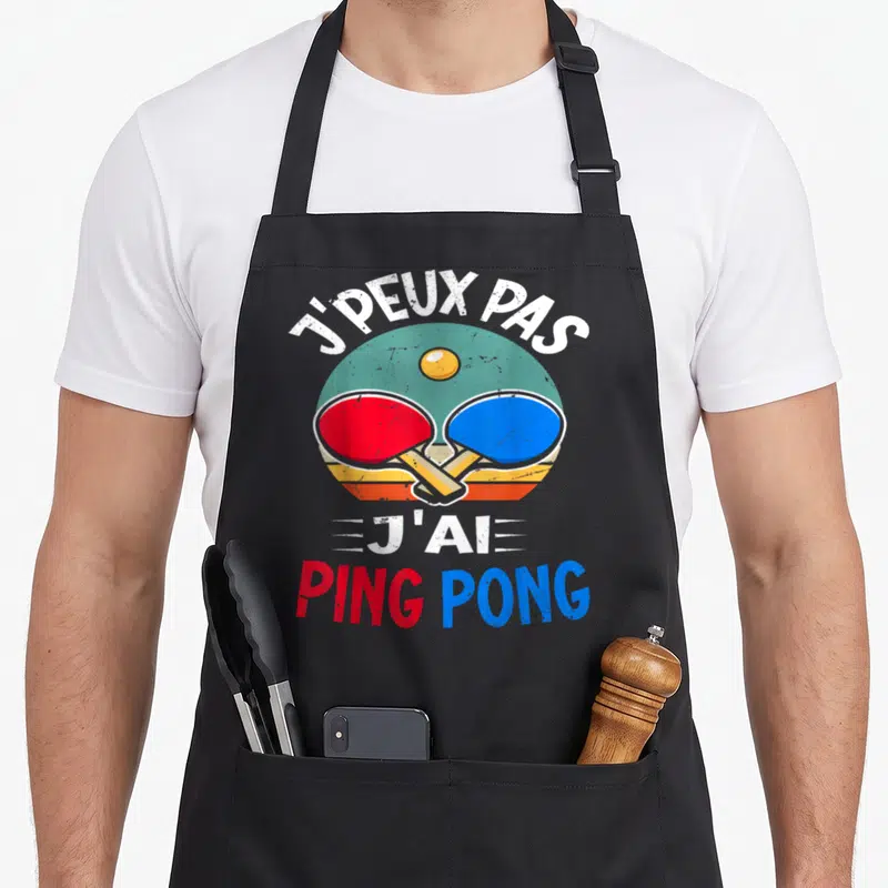 Tablier de cuisine noir avec inscription Je peux pas j'ai ping pong et deux raquettes colorées croisées sur fond circulaire vintage.