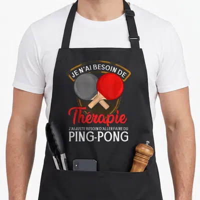 Tablier de cuisine noir avec deux raquettes de ping-pong rouge et noire croisées sous un texte humoristique en blanc et rouge.
