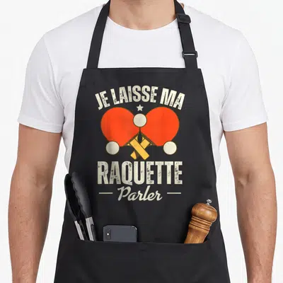 Tablier de cuisine noir avec deux raquettes de ping-pong oranges croisées et le texte Je laisse ma raquette parler en blanc.