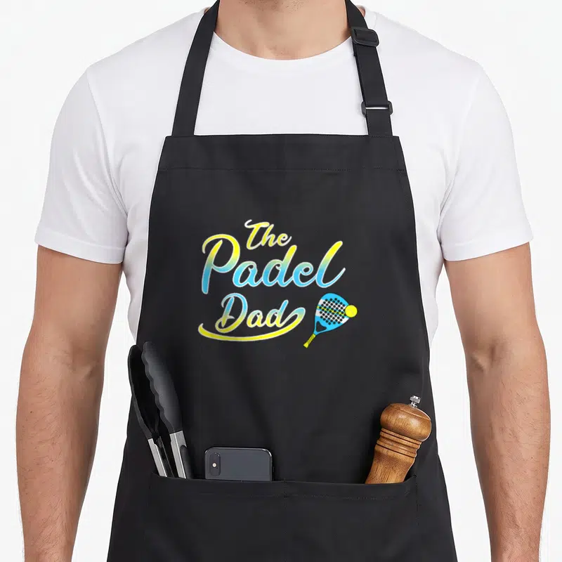 Tablier de cuisine noir avec The Padel Dad en lettres jaunes et bleues accompagnées d'une raquette et d'une balle de sport.