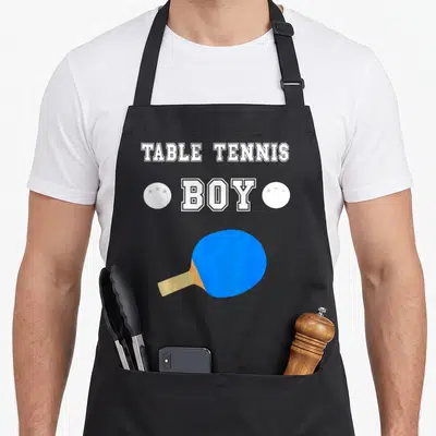 Tablier de cuisine noir avec inscription Table Tennis Boy et raquette de ping-pong bleue sur fond gris uni.