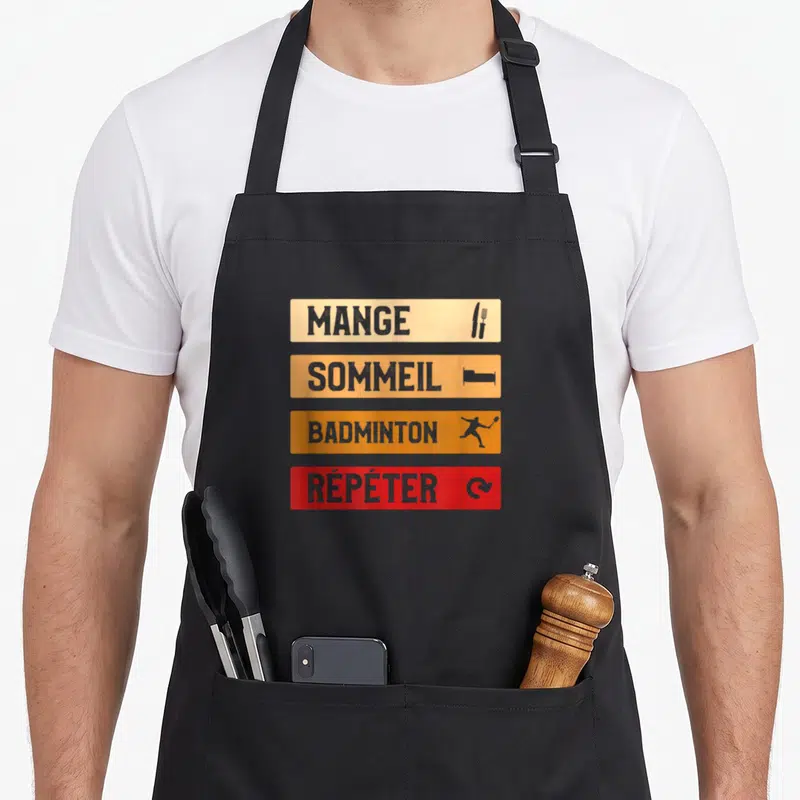 Tablier de cuisine noir avec quatre bandes colorées portant les mots Mange Sommeil Badminton Répéter accompagnés d'icônes.