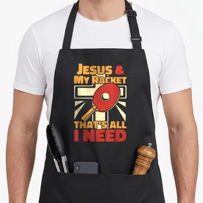 Tablier de cuisine noir avec inscription orange et rouge et illustration de raquette de ping-pong devant une croix stylisée.