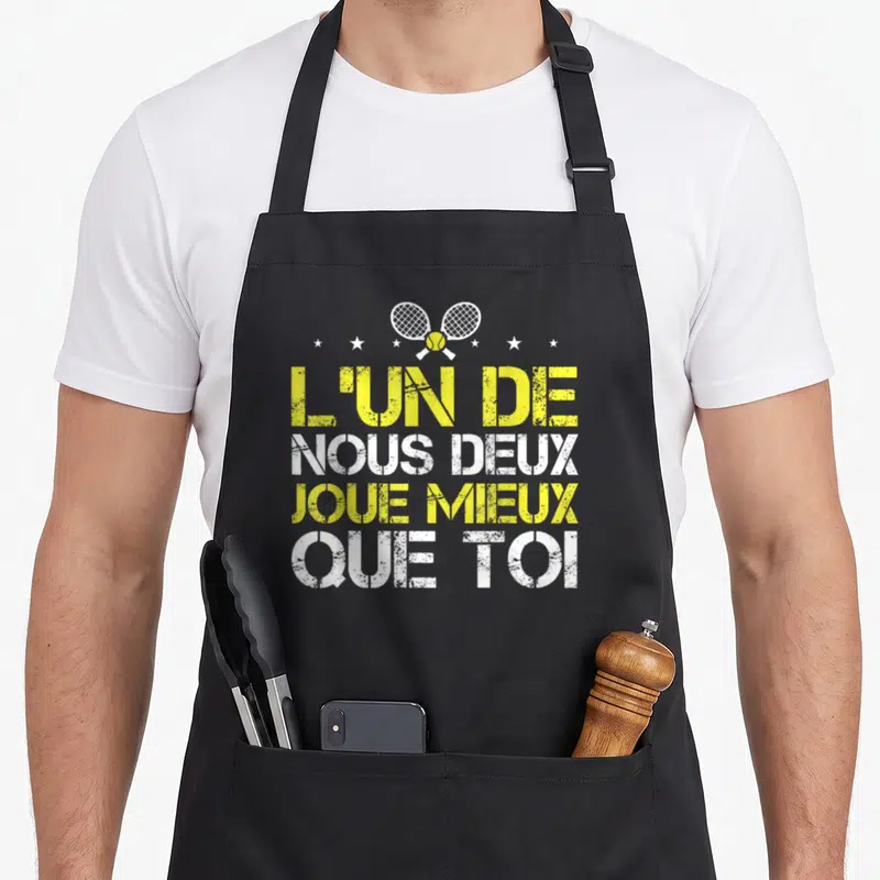 Tablier de cuisine noir avec inscription jaune et blanche L'un de nous deux joue mieux que toi sur fond gris uni