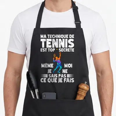 Tablier de cuisine noir avec silhouette multicolore de joueur de tennis et texte blanc humoristique sur fond uni.