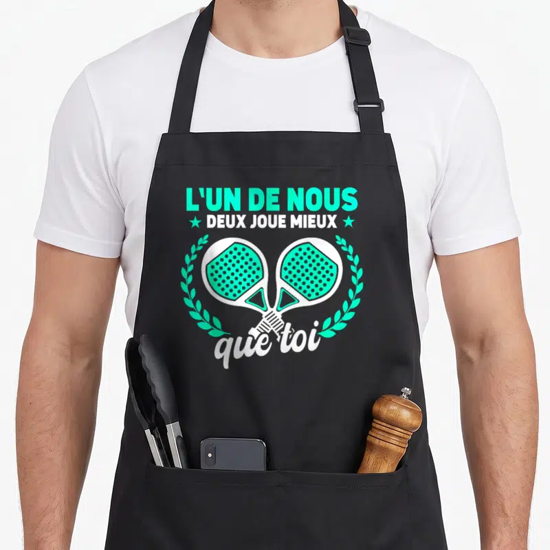 Tablier de cuisine noir avec deux raquettes de padel turquoise croisées et une inscription humoristique en blanc et vert.