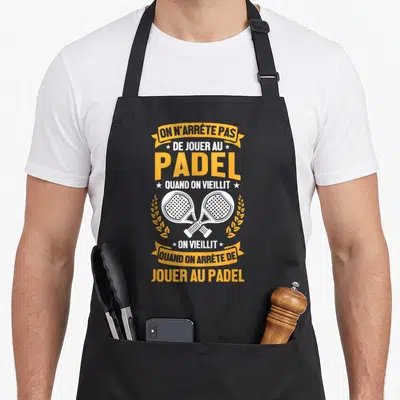 Tablier de cuisine noir avec raquettes de padel croisées et texte jaune sur fond gris uni.