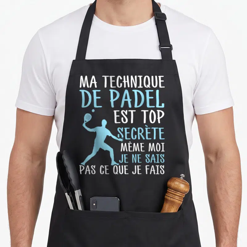 Tablier de cuisine noir avec silhouette bleue d'un joueur de padel et texte blanc et bleu humoristique sur la technique secrète.