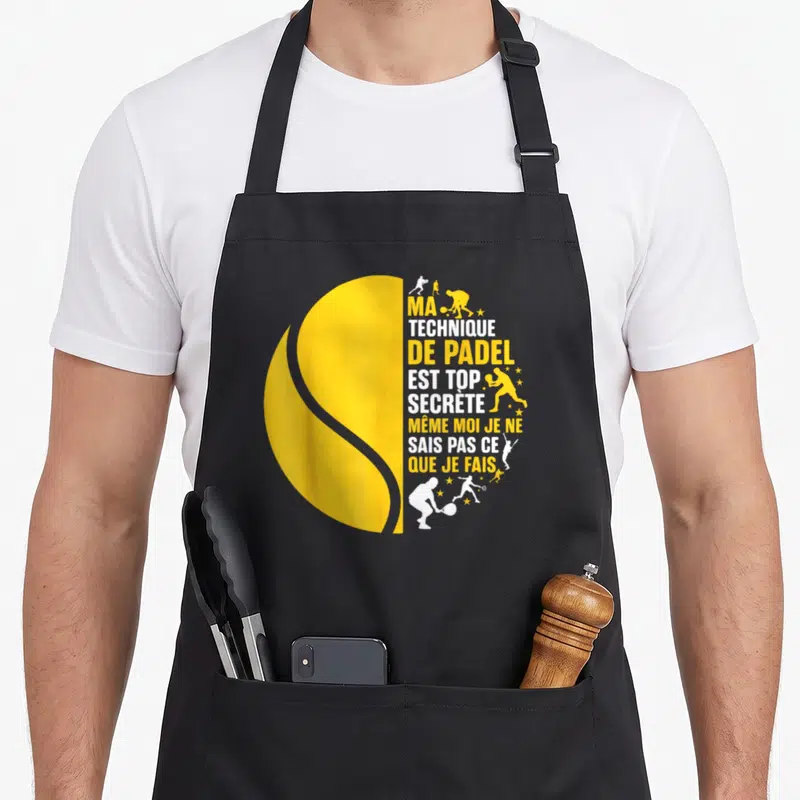 Tablier de cuisine noir avec motif balle de padel jaune et silhouettes de joueurs en action sur fond blanc et orange.