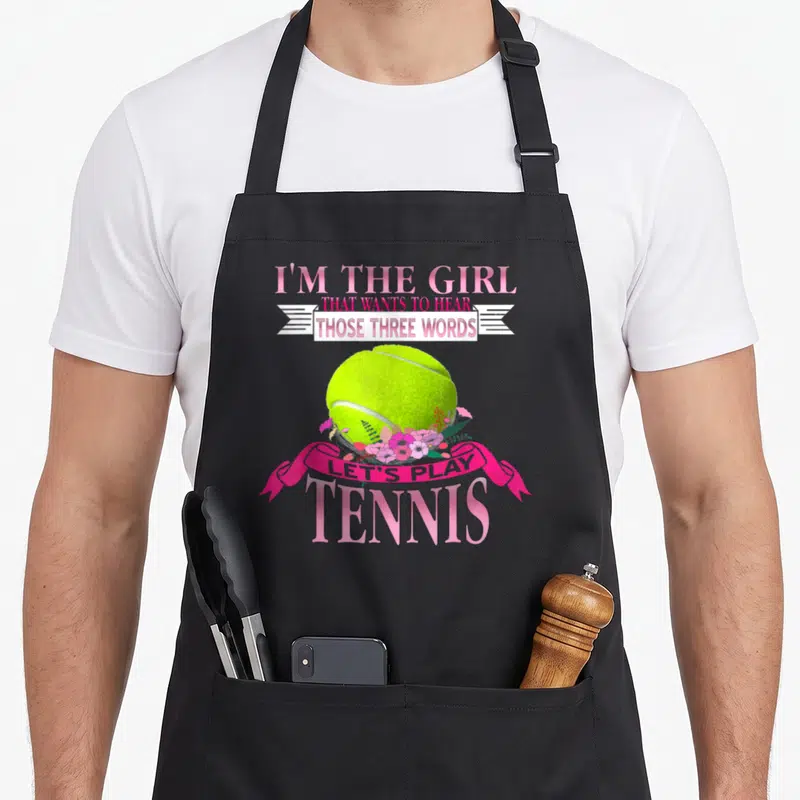 Tablier de cuisine noir avec un ballon de tennis vert, des fleurs roses et le texte I'm the girl that wants to hear those three words.