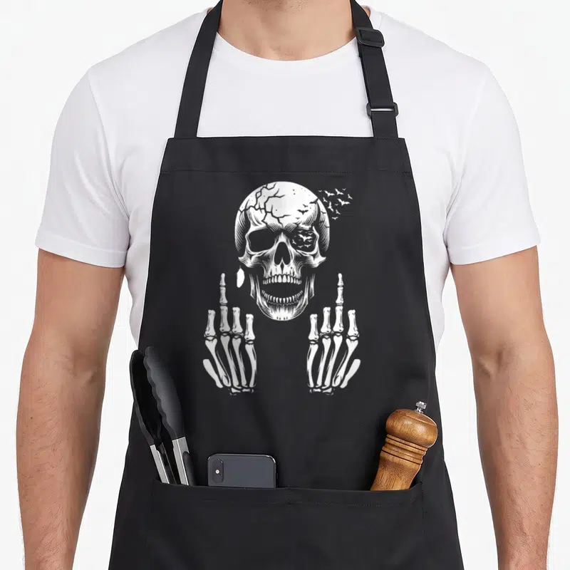 Tablier de cuisine noir avec un crâne et deux mains squelettiques faisant le doigt d'honneur en blanc.
