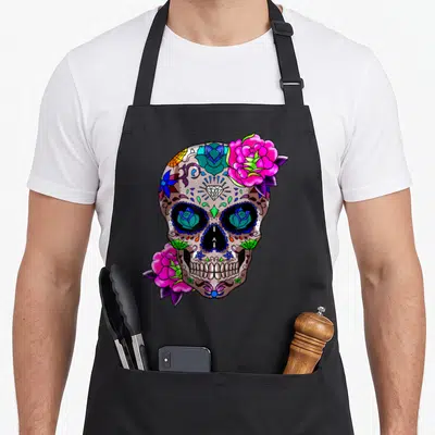 Tablier de cuisine noir avec un crâne décoré de roses roses, de motifs tribaux et de roses bleues dans les orbites.