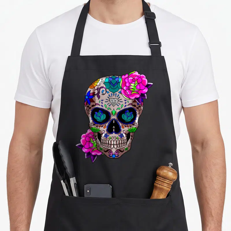 Tablier de cuisine noir avec un crâne décoré de roses roses, de motifs tribaux et de roses bleues dans les orbites.