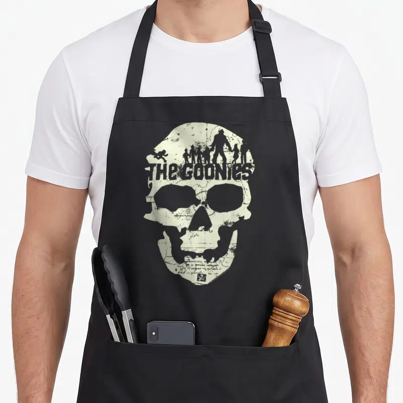 Tablier de cuisine noir avec un grand crâne beige intégrant des silhouettes et le texte The Goonies style carte vieillie.