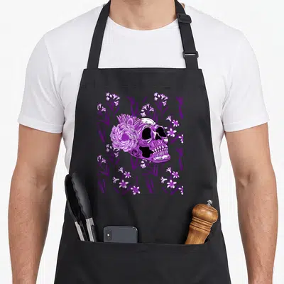 Tablier de cuisine noir avec tête de mort violette entourée de fleurs et motifs floraux sombres.