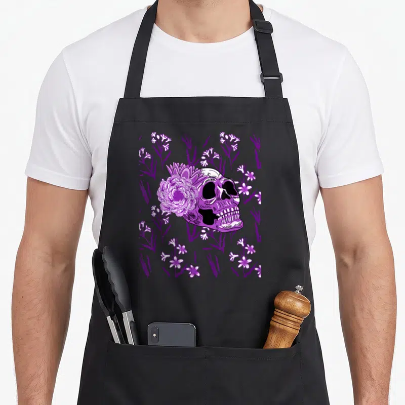 Tablier de cuisine noir avec tête de mort violette entourée de fleurs et motifs floraux sombres.