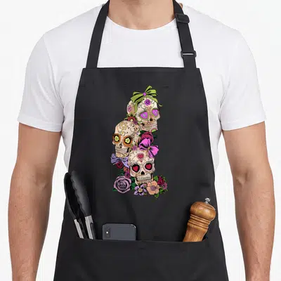 Tablier de cuisine noir avec trois têtes de mort décorées de fleurs roses, violettes et de nœuds colorés dans un style Dia de los muertos.