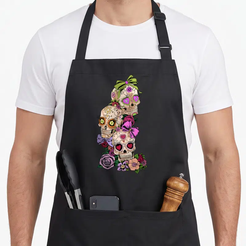 Tablier de cuisine noir avec trois têtes de mort décorées de fleurs roses, violettes et de nœuds colorés dans un style Dia de los muertos.