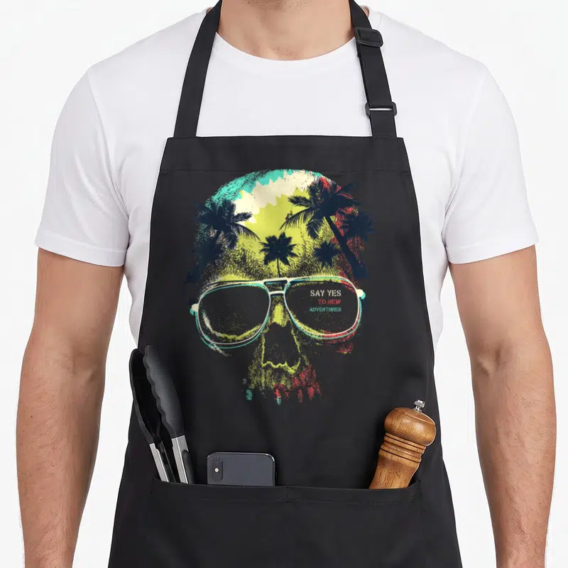 Tablier de cuisine noir avec un crâneSugar Skull portant des lunettes aviateurs sur fond de palmiers colorés.