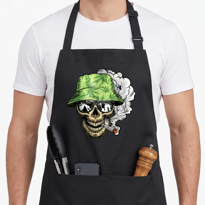 Tablier de cuisine noir avec un crâne fumant un CBD portant un chapeau vert à motifs de feuilles de cannabis et des lunettes de soleil.