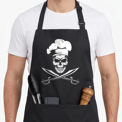 Tablier de cuisine noir avec tête de mort portant un bonnet de chef et deux couteaux croisés.