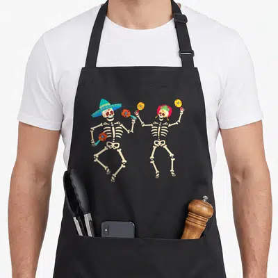 Tablier de cuisine noir avec deux squelettes dansant au Jour des Morts tenant des maracas colorés.