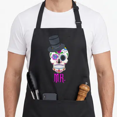 Tablier de cuisine noir avec une calavera colorée portant un chapeau haut de forme et le mot MR en rose sous le crâne.