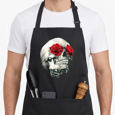 Tablier de cuisine noir avec une tête de mort illustrée portant des roses rouges vives sur les orbites.