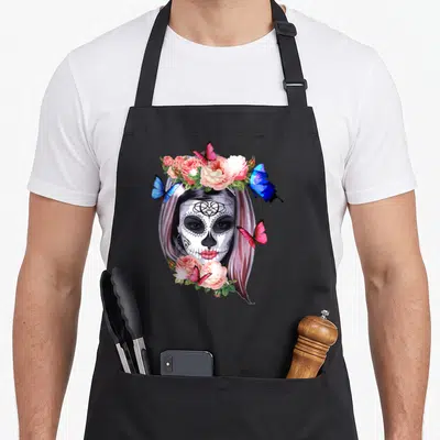Tablier de cuisine noir avec illustration crâne gothic floral, roses roses et papillons bleus et roses sur le devant.