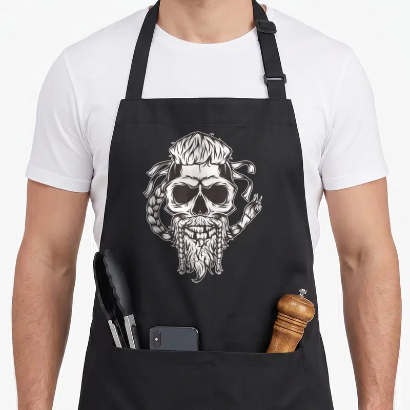 Tablier de cuisine noir avec un crâne viking barbu aux cheveux tressés en dessin blanc.