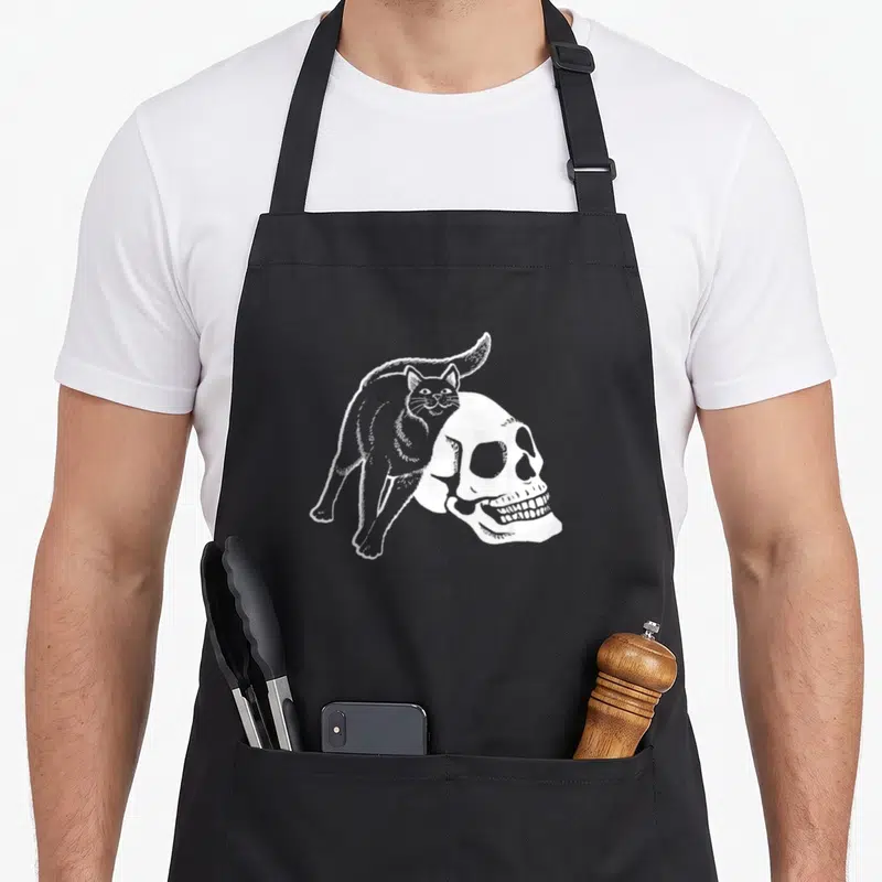 Tablier de cuisine noir avec illustration blanche d'un chat noir posé sur une tête de mort stylisée style gravure.