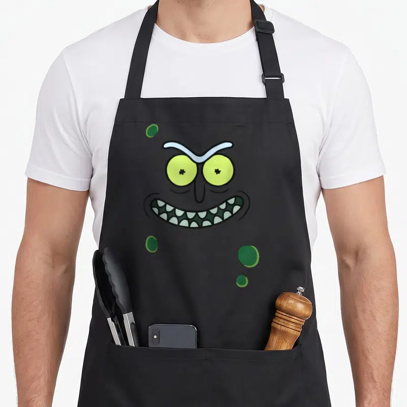 Tablier de cuisine noir avec le visage vert de Pickle Rick, yeux ronds et large sourire aux dents pointues.