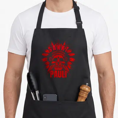 Tablier de cuisine noir avec une tête de mort rouge coiffée de plumes et le texte Sankt Pauli imprimé au centre.
