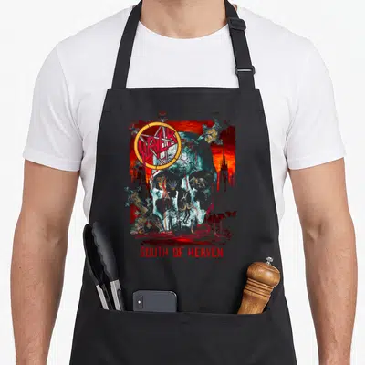 Tablier de cuisine noir avec une tête de mort bleutée, le logo Slayer en cercle orange et l'inscription South of Heaven en bas.