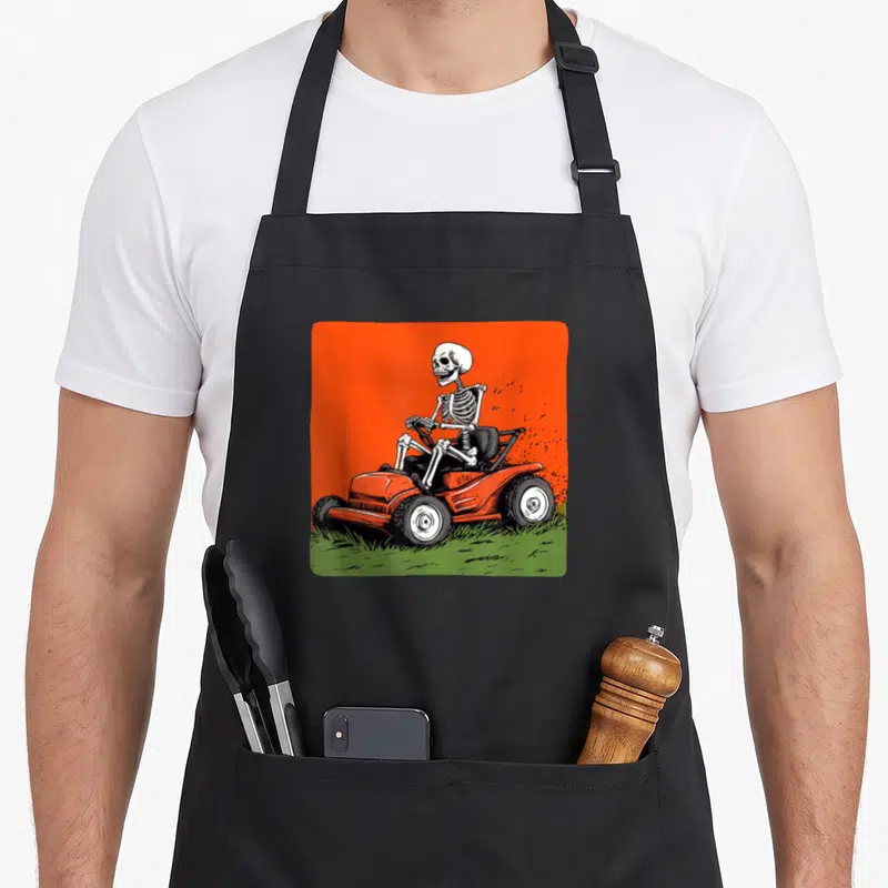 Tablier de cuisine noir avec illustration d'un squelette conduisant une tondeuse à gazon sur fond orange vif.