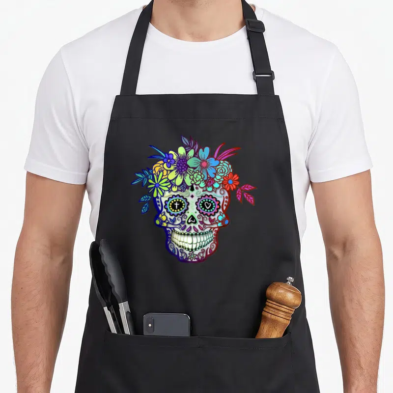 Tablier de cuisine noir avec une tête de mort décorée de fleurs multicolores et des motifs calavera traditionnels.