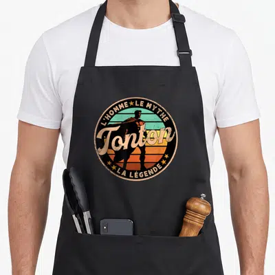 Tablier de cuisine noir avec logo circulaire rétro super tonton lhomme le mythe la légende et silhouette de super-héros sur fond de soleil.