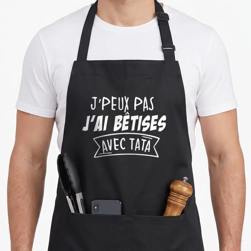 Tablier de cuisine noir avec l'inscription blanche J'peux pas j'ai bêtises avec tata en typographie manuscrite.