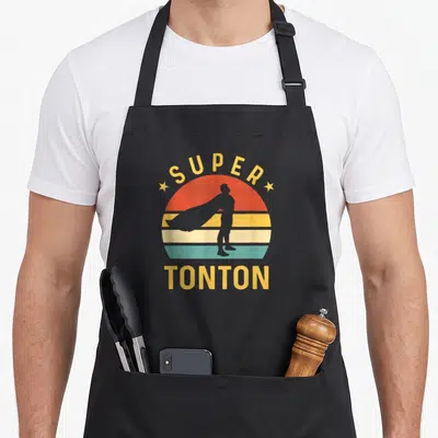 Tablier de cuisine noir avec silhouette de super héros devant un soleil rayé et le texte Super Tonton Meilleur Oncle du Monde.