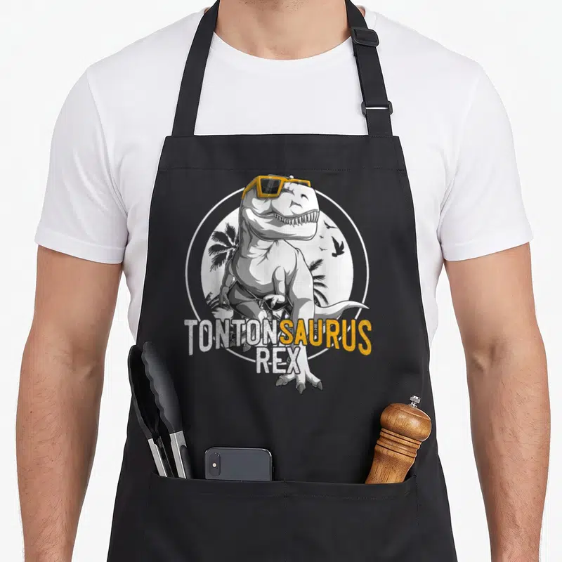 Tablier de cuisine noir avec illustration dinosaure aux lunettes jaunes et texte Tontonsaurus Rex.