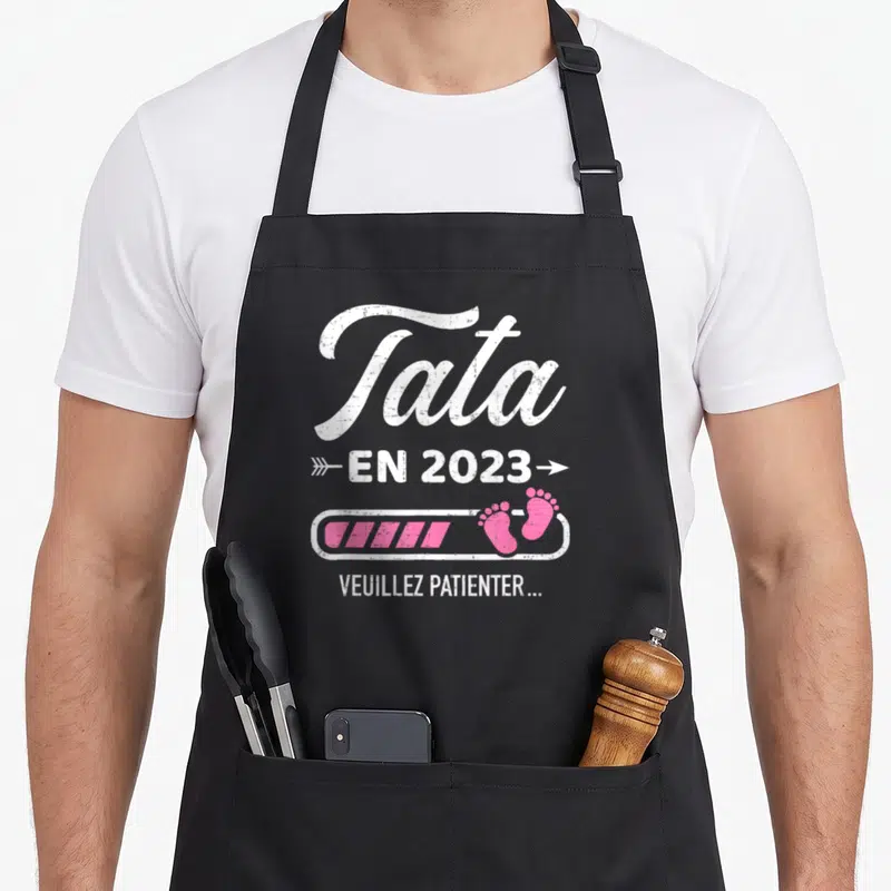 Tablier de cuisine noir avec inscription Tata en 2023 veuillez patienter et petits pas roses sur une barre de progression.