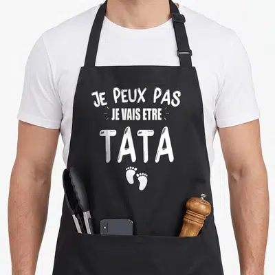 Tablier de cuisine noir avec le texte Je peux pas je vais être tata et deux petits prints de pieds de bébé en dessous.