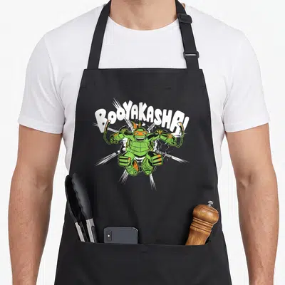 Tablier de cuisine noir avec illustration colorée de Mikey brandissant des nunchaks sous le texte Booyakasha.