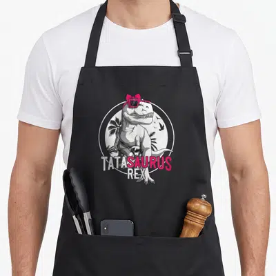 Tablier de cuisine noir avec un dinosaure portant des lunettes roses et un nœud, entouré du texte TATASAURUS REX.