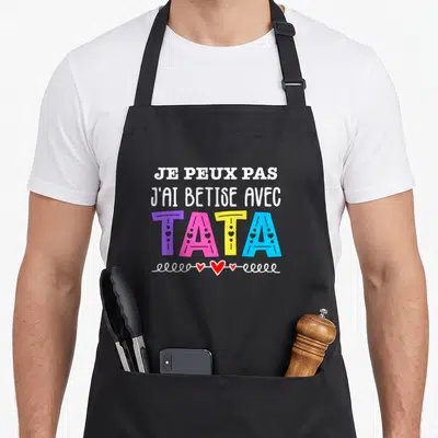 Tablier de cuisine noir avec l'inscription Je peux pas j'ai bêtise avec Tata en lettres multicolores ornées de petits cœurs rouges et blancs.