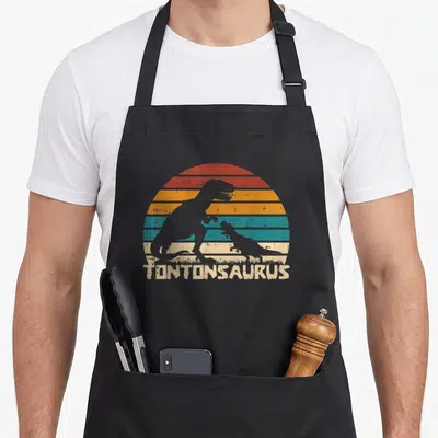Tablier de cuisine noir avec silhouette de dinosaure et texte Tontonsaurus sur fond de soleil rayé vintage
